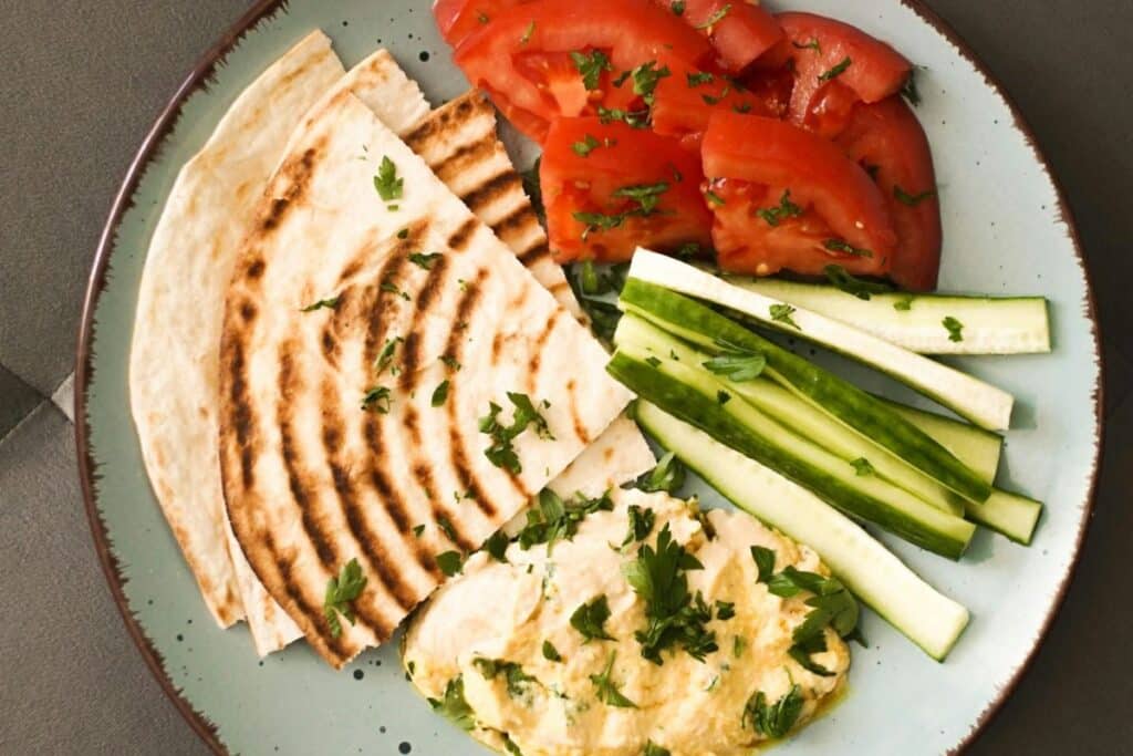 plate full of plant based snacks including pitas, tomatoes, zuchinni and hummus